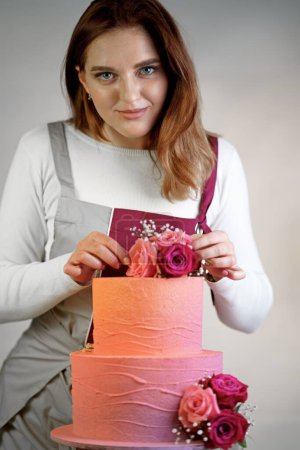 Téléchargez les photos : Confiseur dans un tablier de travail décore un gâteau d'anniversaire avec des fleurs fraîches - en image libre de droit