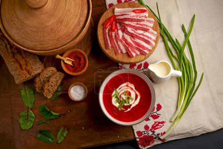 Traditional Ukrainian borsht, red vegetable soup or borscht with smetana on wooden background. Slavic dish with cabbage, beets, tomatoes Traditional Ukrainian towel along with garlic, bread and salt. Top view of a wooden tray on a black background on