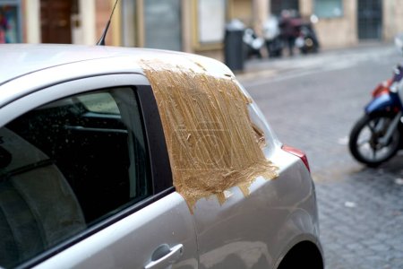 A car with a makeshift cover on its broken window, an improvised solution amidst the urban backdrop of city life.