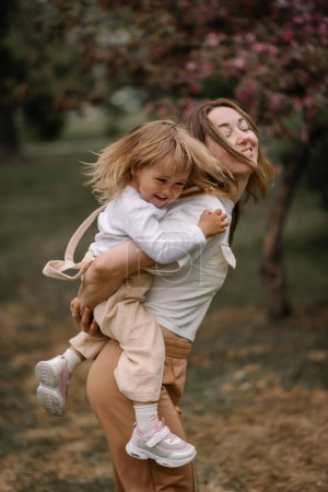 Schöne Mutter und Tochter vor dem Hintergrund eines blühenden Apfelbaums. Mutter reitet ihre kleine Tochter auf dem Rücken. Stilvolle Kleidung in neutralen Farben. Mutter und tochter haben spaß