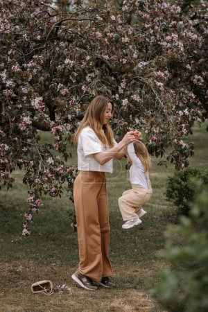 Schöne Mutter und Tochter vor dem Hintergrund eines blühenden Apfelbaums. Mama hält ihre kleine Tochter an den Händen. Stilvolle Kleidung in neutralen Farben. Mutter und tochter haben spaß