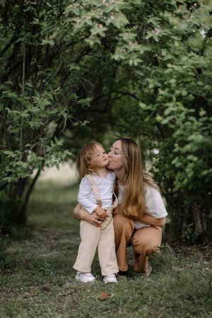 Ein kleines stilvolles Mädchen, ein Kind und ihre Mutter setzten sich auf einen grünen Rasen in einem Park zwischen Büschen. Mutter umarmt ihre Tochter. Die Tochter hält einen Korb und einen Zweig eines blühenden Apfelbaums in der Hand.