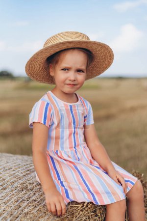 Téléchargez les photos : Une petite fille en été, portant une robe à rayures multicolores et un chapeau de paille, s'assoit sur une balle de paille et regarde au loin. Fond flou - en image libre de droit