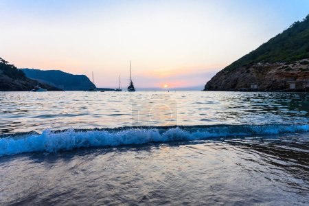 Foto de Hermosa puesta de sol en la playa de Benirras en la parte noroeste de Ibiza, España. Hay olas pequeñas en la playa. La formación rocosa de Cap Bernat, también conocido como Dedo de los Dioses, se puede ver en el fondo - Imagen libre de derechos
