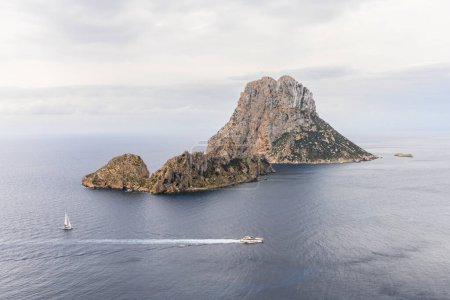 Foto de Islas características de Es Vedra y Es Vedranell frente a la costa sur de Ibiza, vistas desde la meseta Mirador des Vedra, en un día nublado. Un velero y un yate pasan por la isla. Es Vedra es el tercer punto más magnético de la Tierra. - Imagen libre de derechos