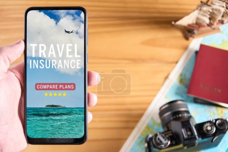 Top view of male hand holding mobile phone with travel insurance website on the screen. Travel items in the background with camera, passport and map. Travel insurance website or app concept. Copy space.