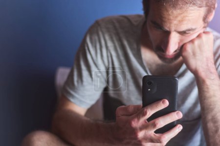 Téléchargez les photos : Jeune homme caucasien de 30 ans assis à la maison tout en regardant l'écran de téléphone portable avec ennuyé, expression désintéressée contre le mur bleu. Ennuyeux, dépendance à la technologie, isolement et dépression. - en image libre de droit