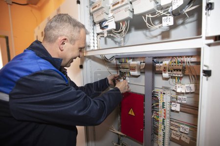 An electrician in overalls checks a shield with electric plugs and wires.
