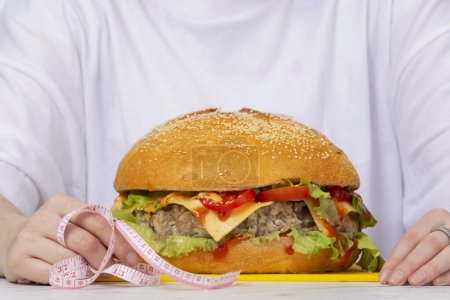 Photo for Big burger measuring tape on the table against the background of female hands. The concept of diet and elimination of excess calories. - Royalty Free Image