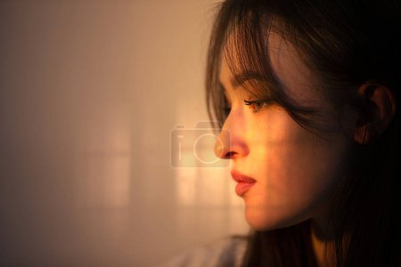 Photo for The face of a pensive girl in profile is illuminated by warm light from the window. - Royalty Free Image