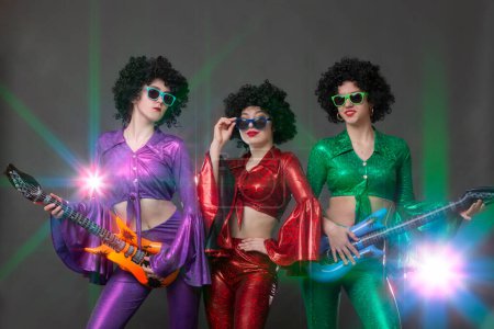 Photo for Beautiful girls dressed in disco style in afro wigs pose with guitars in the rays of stage light. Vintage disco group for women. - Royalty Free Image