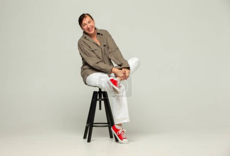 Middle-aged woman in casual clothes and red sneakers poses with a smile in the studio on a gray background