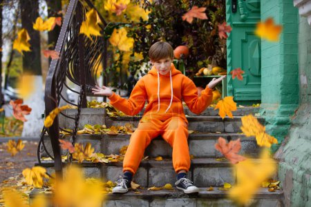 A boy in orange clothes sits on the steps and looks at the yellow flying autumn leaves. Fall has come.