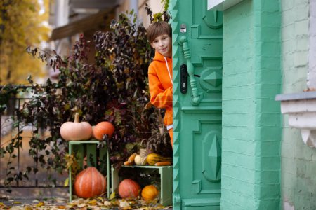 Un garçon en vêtements orange regarde par la porte d'une maison décorée de citrouilles pour Halloween.