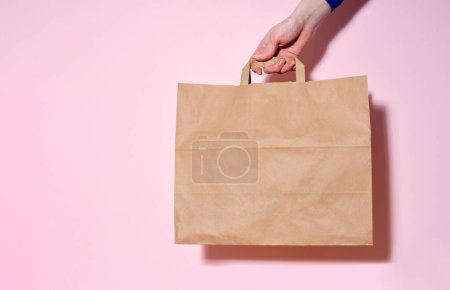 Téléchargez les photos : Main féminine tenant un sac en papier avec des articles livrés. Fond rose, espace de copie - en image libre de droit