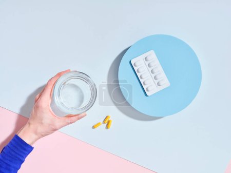 Foto de Composición creativa con píldoras de suplemento alimenticio, vaso de agua y mano femenina sobre fondo colorido - Imagen libre de derechos