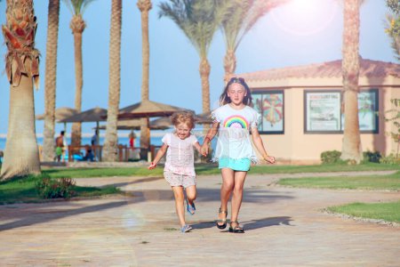 Happy sisters run on footpath in tropical resort. Children in palm park. Joyfull girls run on path among date trees. Children enjoying holidays. Sisters on vacations. Girls relaxing