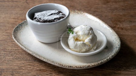 Photo for Molten chocolate lava cake and a scoop of ice cream served in ceramic jars, on a white plate with a silver spoon, on the wooden table, close-up shot. - Royalty Free Image
