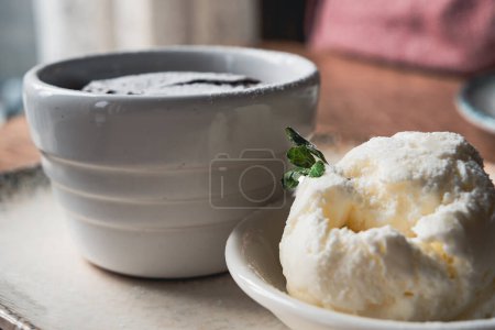 Photo for Molten chocolate lava cake and a scoop of ice cream served in ceramic jars, on a white plate with a silver spoon, on the wooden table, close-up shot. - Royalty Free Image