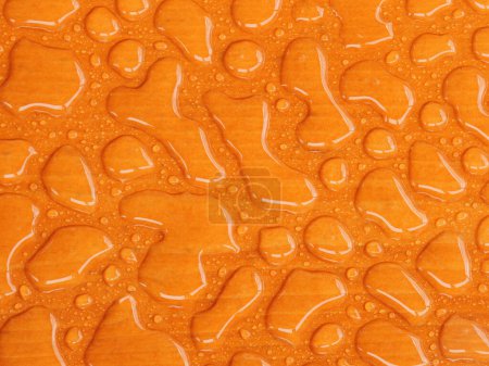 Water droplets forming a unique texture on a smooth, varnished orange color wooden surface, highlighting the interaction between water and wood.