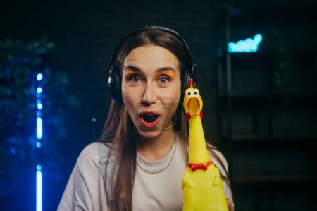 Photo for Funny girl in headphones with a rubber chicken in her hands with a shocked face looking at the camera. - Royalty Free Image