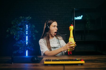 Photo for Shocked gamer girl sits at the table with a rubber chicken toy in her hands and looks at it with a shocked face while playing online. - Royalty Free Image