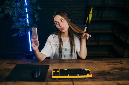 Photo for Positive female gamer sits at home at the table and plays games, takes a selfie on the smartphone camera - Royalty Free Image