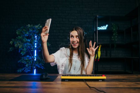 Photo for Positive gamer girl plays online games and streams on computer and records video for followers with a smile on her face on smartphone camera. - Royalty Free Image