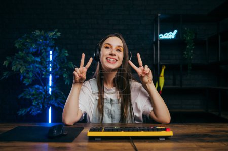 Photo for Cute female gamer wearing headphones showing peace gesture and looking at camera with smile on her face while streaming game - Royalty Free Image