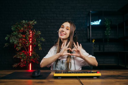 Photo for Positive female gamer in headphones sitting at the table at home while playing online game on the computer and showing a heart gesture, looking at the camera and smiling. - Royalty Free Image