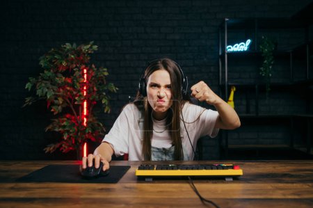 Photo for Angry female gamer in casual clothes sitting at the table and playing games on the computer at home, showing her fist to the camera and grimacing. - Royalty Free Image