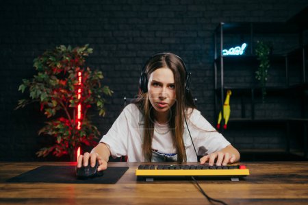 Photo for Concentrated female gamer in a headset sits at a table with a serious face and plays online games with a team on a computer. - Royalty Free Image