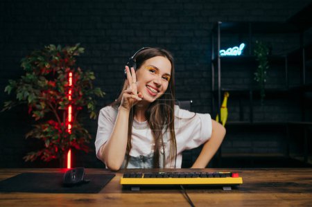 Photo for Cheerful gamer girl sits at the table in a headset and with a smile on her face shows a peace gesture and looks at the camera, plays online games at home. - Royalty Free Image