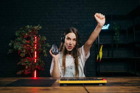 Photo for Attractive cheerful woman in headphones with a mouse in her hands rejoices at the victory in the game on the computer, looks at the camera with a joyful face. - Royalty Free Image