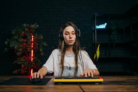 Photo for Beautiful female gamer in a headset sits at home at the table playing online games on the computer with a calm face. - Royalty Free Image