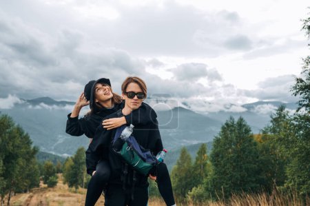 Photo for Happy woman and man hikers in stylish casual clothes having fun in the mountains, the guy lifted his girlfriend on his back. - Royalty Free Image