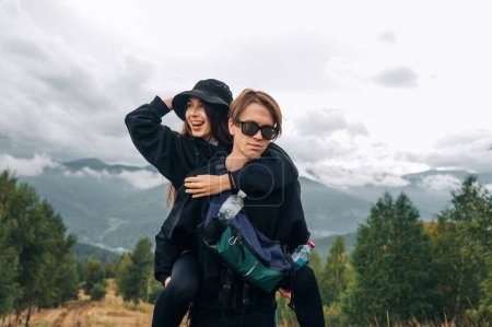 Photo for A stylish couple of cheerful young tourists are having fun on the top of a mountain, a woman is smiling, a man is carrying her on his back, standing against the background of beautiful mountain views. - Royalty Free Image