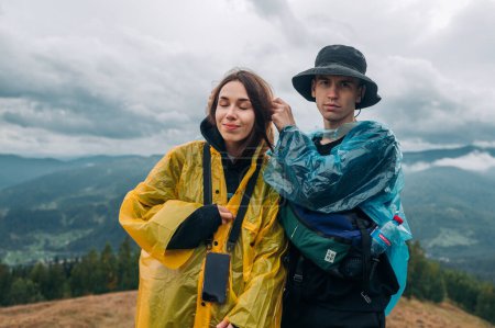 Photo for A cute couple of tourists on a hike in the mountains wearing raincoats pose for the camera against the background of beautiful views. The couple spends time together on a trip to the mountains - Royalty Free Image