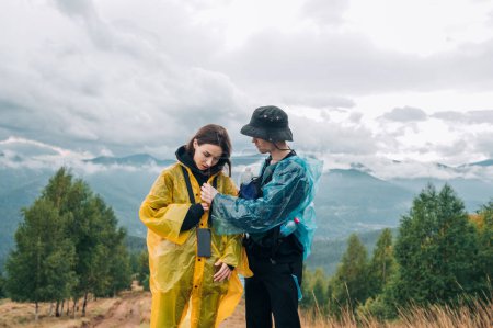 Photo for Young couple of tourists man and woman in the rain in the mountains stand on the background of beautiful views, dressed in casual clothes and raincoats. - Royalty Free Image