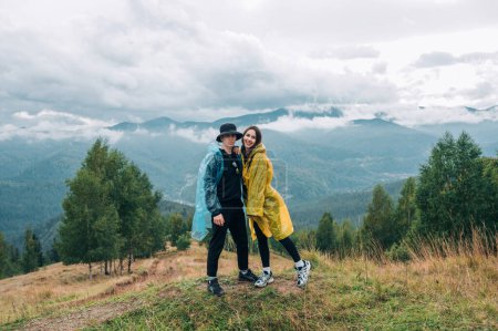 Photo for Beautiful young couple man and woman in raincoats while hiking in the mountains posing for the camera against the background of beautiful views with a smile on their faces. Recreation - Royalty Free Image