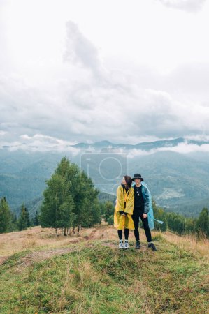 Photo for A young beautiful couple of tourists in raincoats during a hike stand on a mountain against the background of overcast views and pose for the camera, full-length photo. - Royalty Free Image