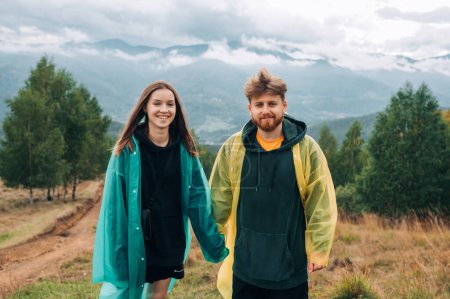 Photo for Photo of a positive beautiful couple in rainy weather in the mountains standing during a hike and posing for the camera against the background of cloudy mountain views. - Royalty Free Image