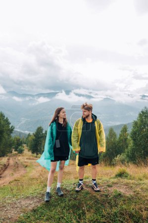 Photo for Beautiful couple man and woman in casual clothes and raincoats standing in the mountains during a hike holding hands and looking at each other with a smile on their face. - Royalty Free Image