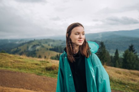 Photo for Positive female tourist in a raincoat stands in the mountains with a smile on her face and looks away. - Royalty Free Image