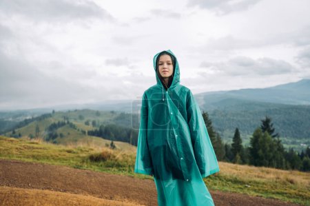 Photo for Attractive woman hiker in raincoat stands in mountains in bad weather during a hike and looks at the camera against the background of beautiful views. - Royalty Free Image