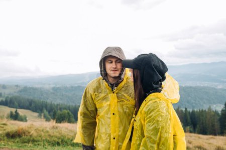 Photo for A beautiful young couple on a hike stands in the mountains against the background of beautiful views, dressed in raincoats and looking at each other with a serious face. - Royalty Free Image