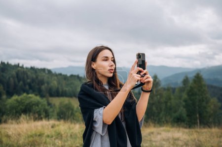 Photo for A beautiful female tourist is standing on a pasture during a hike in the mountains and using a smartphone against the background of beautiful Carpathian views. - Royalty Free Image