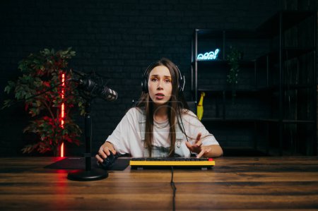 Photo for Shocked female gamer in a headset sits at a computer with a microphone, plays games and looks at the camera with a surprised face. - Royalty Free Image