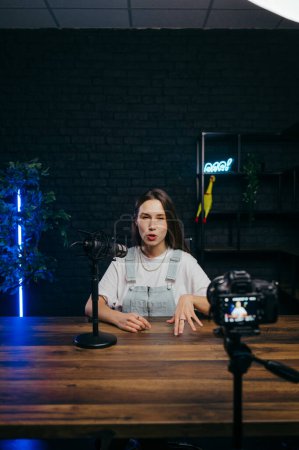 Photo for Funny woman sitting at a table and recording videos on camera with a funny face in a professional studio. Vertical - Royalty Free Image