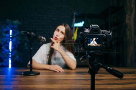 Photo for Funny woman sitting at a table and recording videos on camera with a funny face in a professional studio. Focus on the camera. - Royalty Free Image